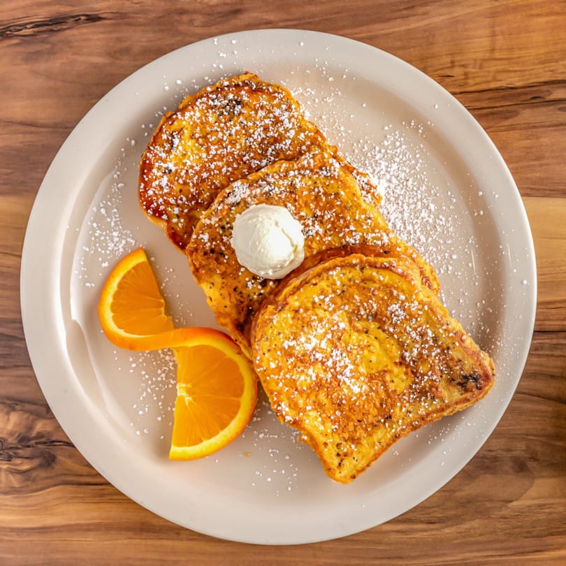 Three slices of golden French toast with powdered sugar, a dollop of butter, and orange slices.