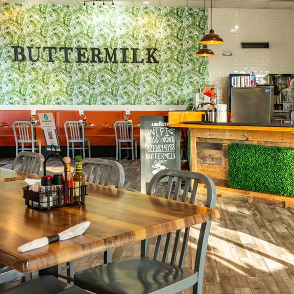 Buttermilk Eatery's interior with a tropical leaf wall, orange booths, and wooden tables with metal chairs.