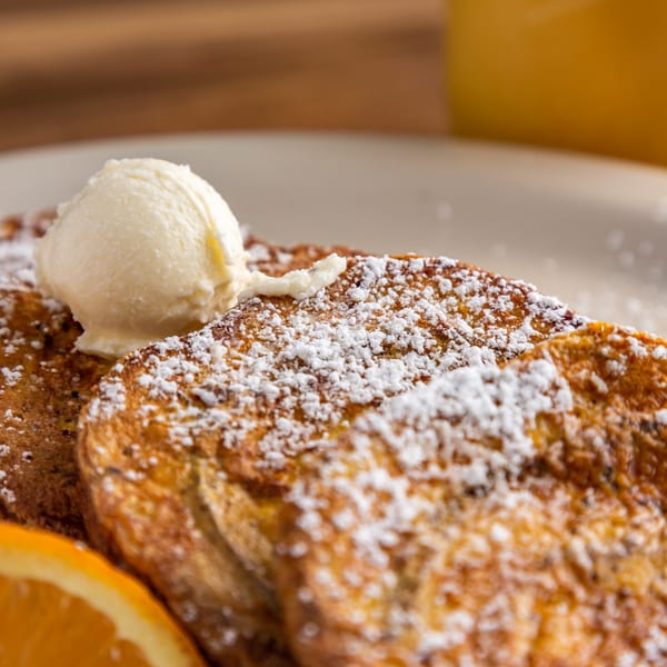 Buttermilk Eatery's French toast, powdered sugar, butter, and an orange slice on a white plate.