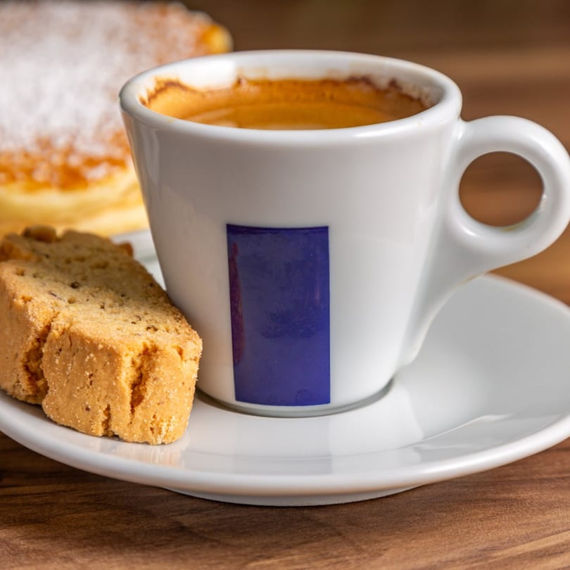 Espresso in a white cup with a blue label, biscotti, and a powdered sugar pastry.