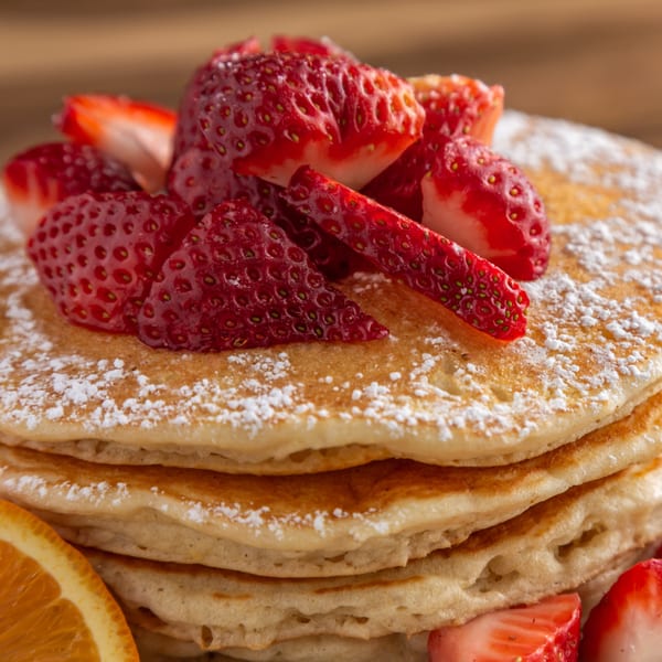 Stack of Buttermilk Eatery pancakes topped with fresh strawberries and powdered sugar, with an orange slice.