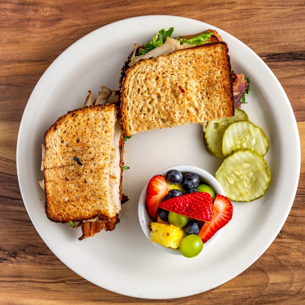Buttermilk Eatery's sandwich with toasted bread, sliced pickles, and a side of fresh fruit.